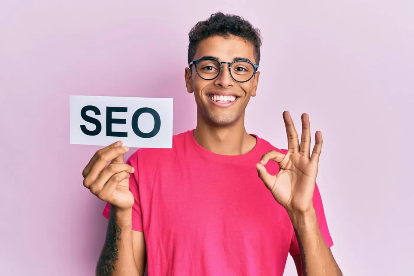 Lächelnde Person mit Brille hält ein Schild mit der Aufschrift „SEO“ und zeigt ein Okay-Zeichen vor hellem Hintergrund.
