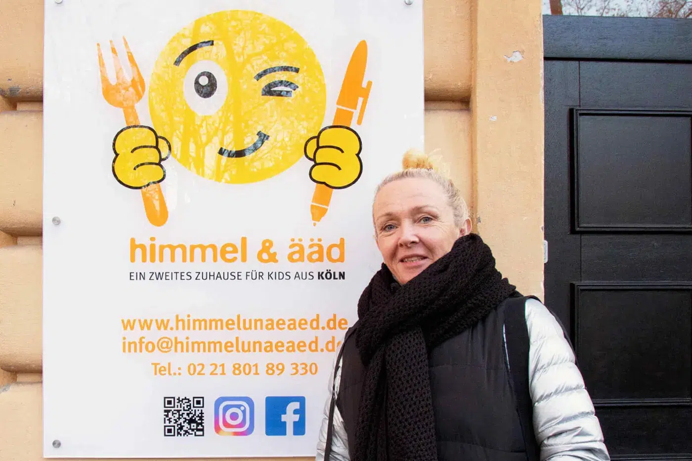 Gabriele Gérard-Post, Geschäftsführerin der gemeinnützigen GmbH himmel & ääd, steht vor einem Schild mit Logo und Informationen ihrer Organisation in Köln.