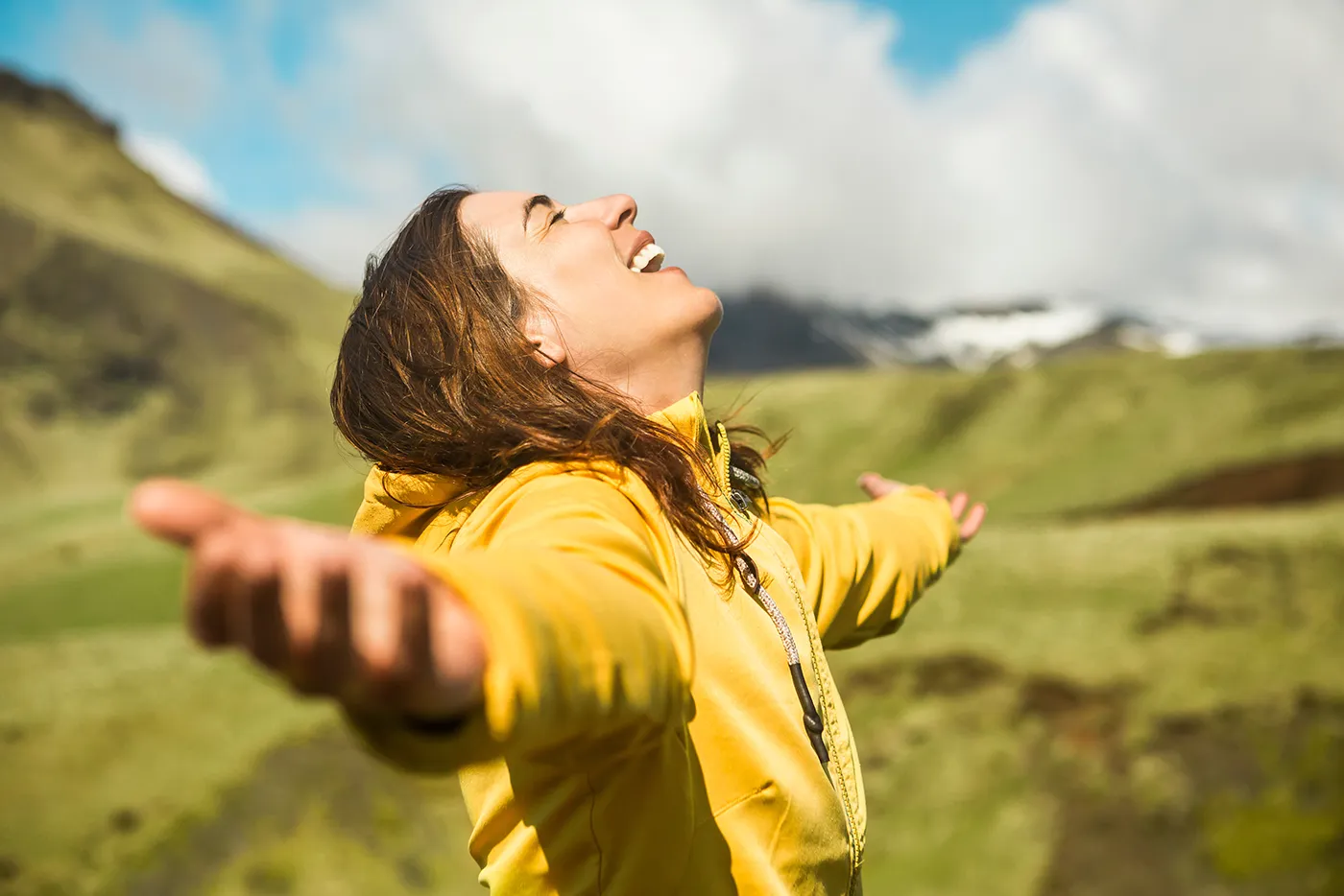 Frau in gelber Jacke steht mit ausgebreiteten Armen in einer grünen Berglandschaft und blickt lächelnd in den Himmel.