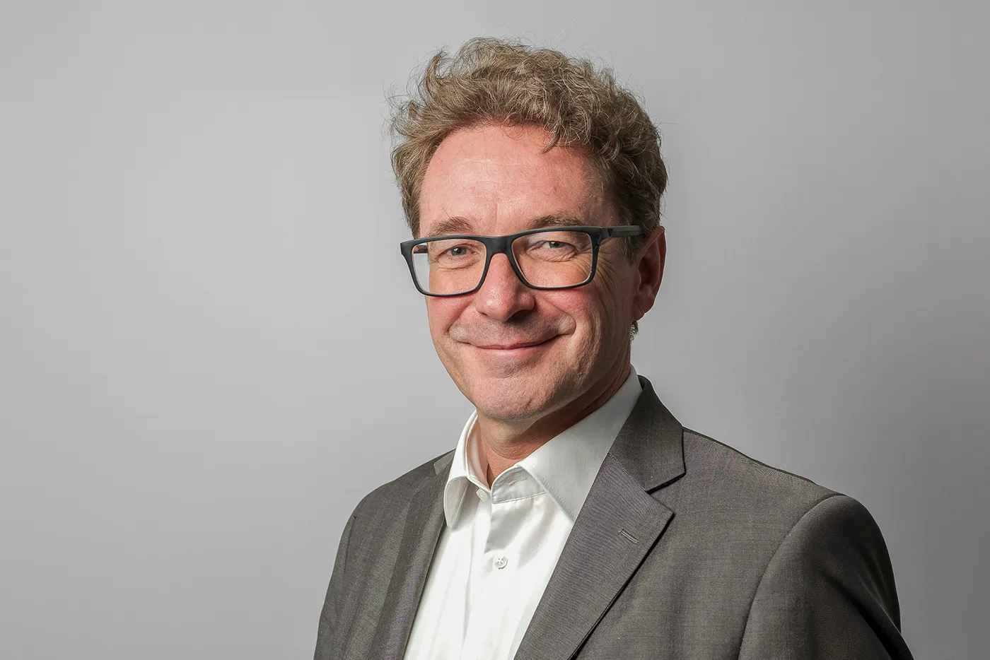 Porträt von Dr. Hamann vor einem hellgrauen Hintergrund. Er trägt eine dunkle Brille, ein weißes Hemd und ein graues Sakko und blickt mit einem leichten Lächeln in die Kamera.