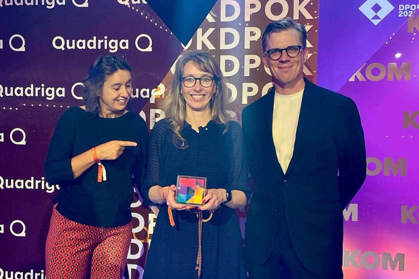 Sabine Haas von der RESULT GmbH (links), Sandra Bulling von der Welthungerhilfe (Mitte) und Jonny Böhm von der Ketchum GmbH (rechts) stehen gemeinsam auf der Bühne einer Preisverleihung. Sandra Bulling hält die DPOK-Auszeichnung in den Händen, während Sabine Haas lächelnd auf sie zeigt. Im Hintergrund sind die Logos von Quadriga und DPOK zu sehen.