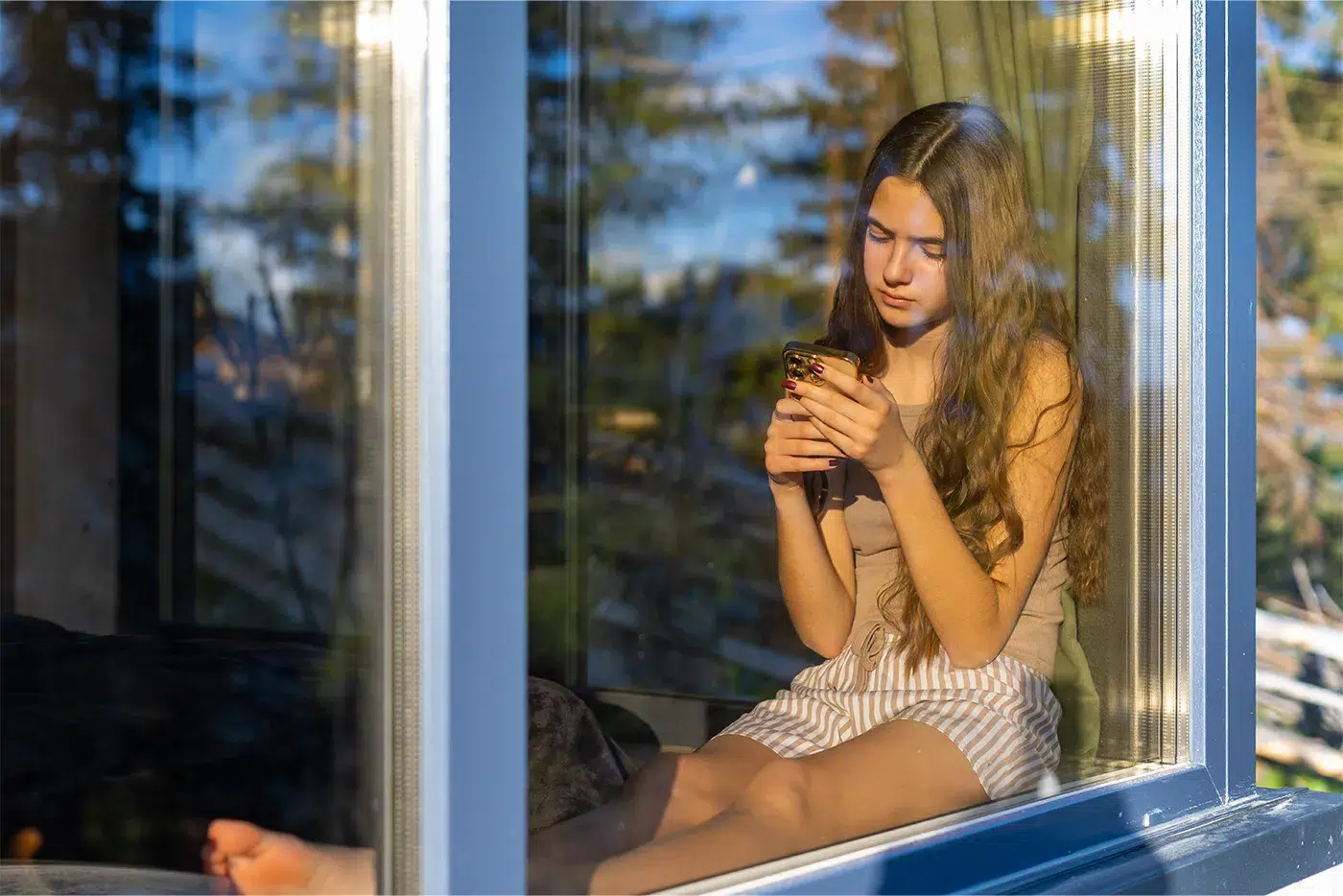 Junge Frau sitzt allein am Fenster und ist mit ihrem Smartphone im Internet.